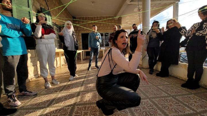 A facilitator is squatting with a lifted pointing finger in the middle of a group of participants of the communication training in Andakit. The participants are holding a yarn that spans a net, connecting them.